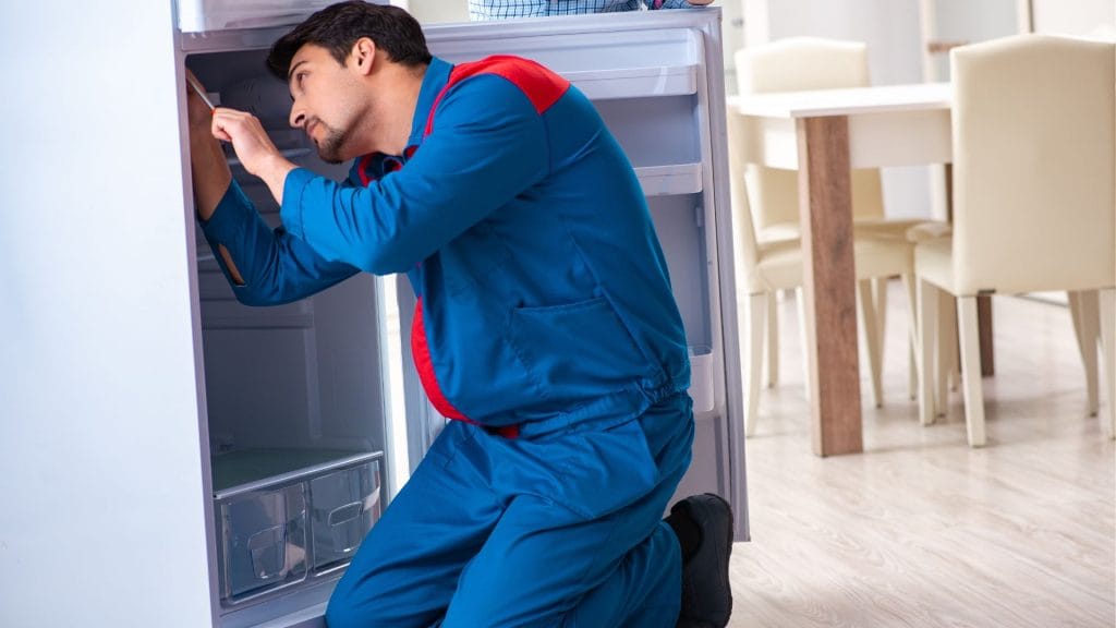 An appliance repair expert in blue overalls repairs a refrigerator, focused on fixing its internal components.