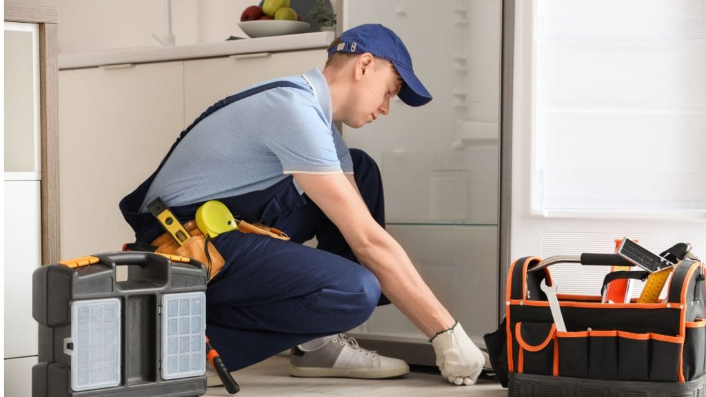 A man in overalls and work boots repairs a refrigerator, focused on his task in a well-lit Ottawa workshop.