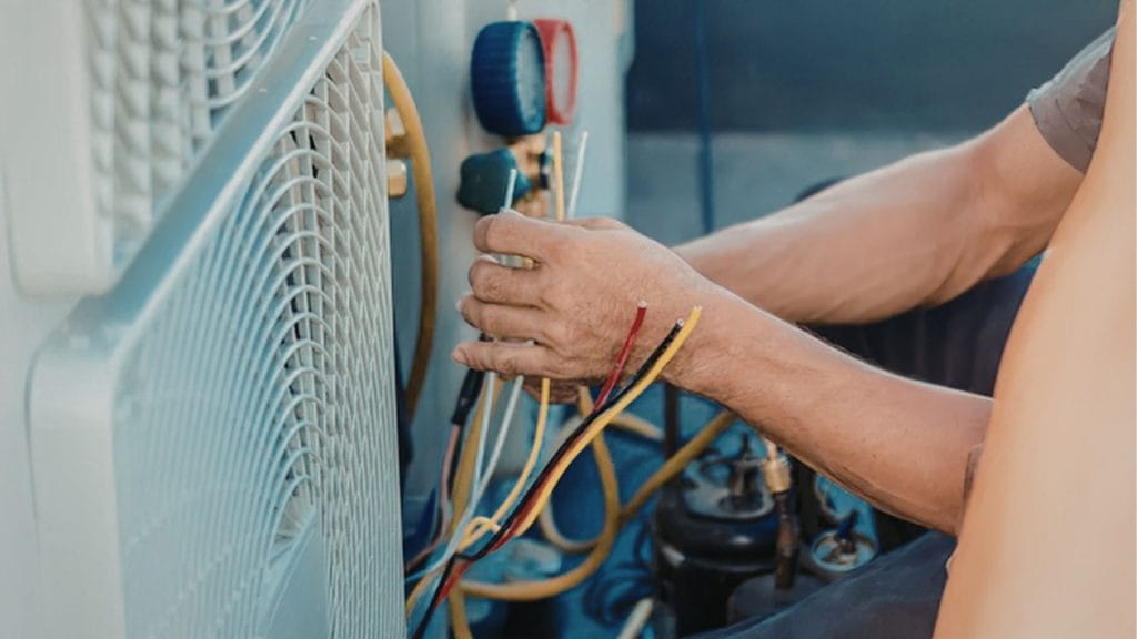 A man repairs an refrigerator, focused on the unit with tools in hand, in a well-lit indoor setting.